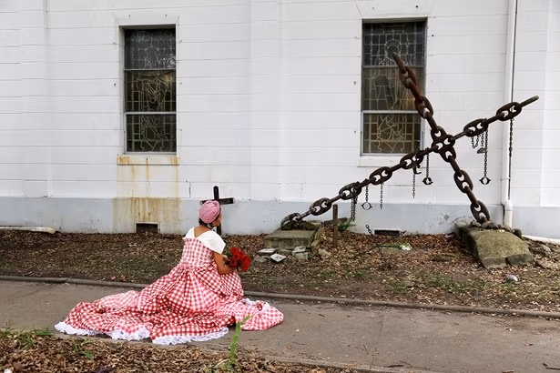 A woman resting in a red and white checkered dress facing a white building. She is holding a bouquet of red flowers.
