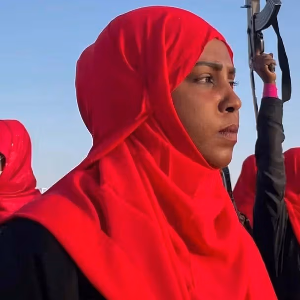 A group of women in bright red headscarves stand outdoors beneath a clear blue sky during a gathering, while a person in the background raises a rifle.