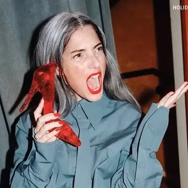 A person sitting backward on a red chair, holding a red high heel to her ear, laughing. Wears a blue shirt and black stockings, with gray hair and red lipstick.