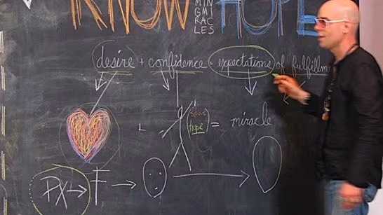 A man at a chalk board in the midst of circling a word. The chalk board has words and diagrams in multiple colors.