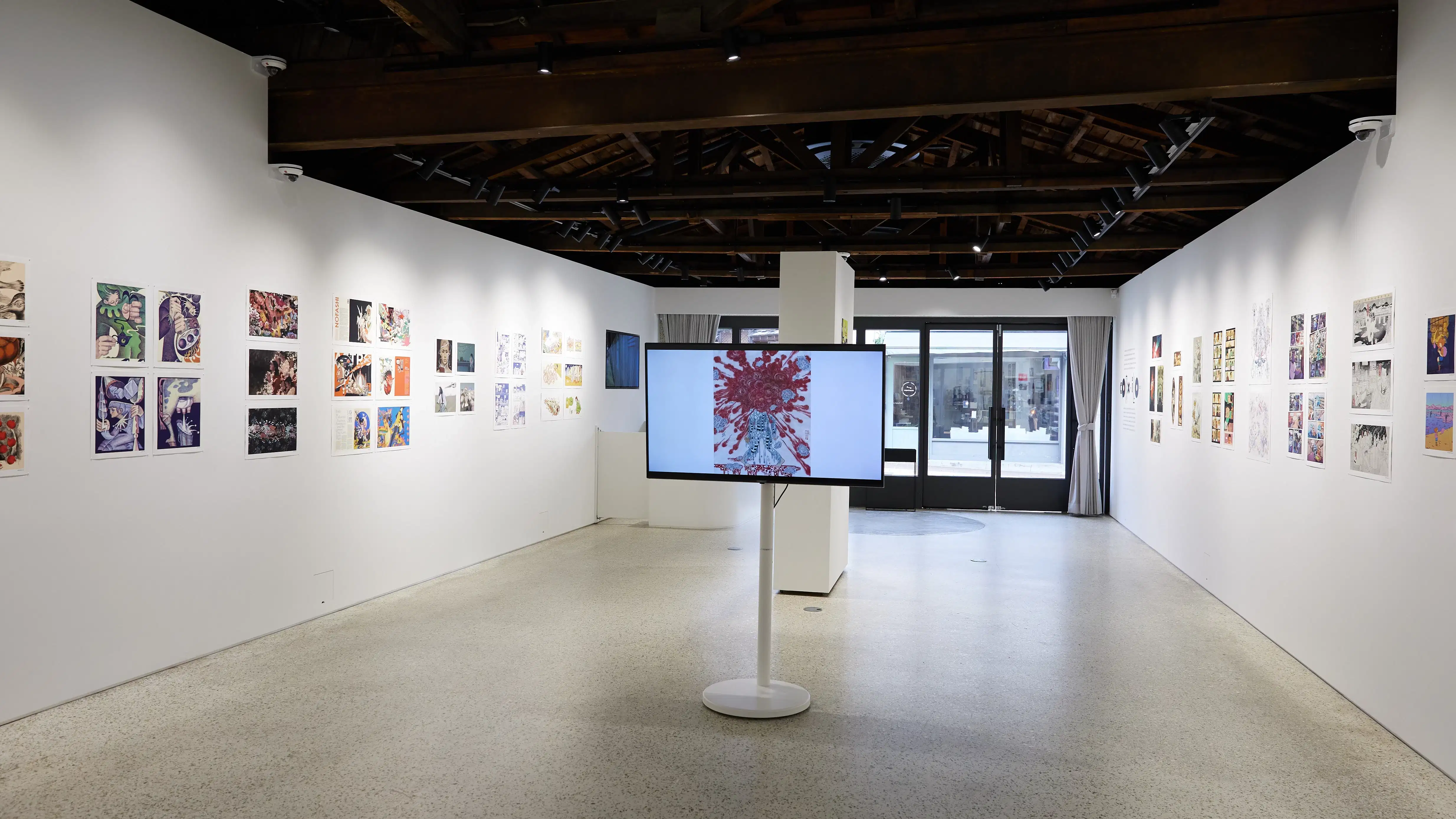 Gallery installation displaying colorful comic and illustration-style artworks arranged linearly along both walls. A large monitor on a white pedestal at the center shows a figure with red abstract patterns. The space features an open ceiling with exposed dark wooden beams, track lighting, and natural light filtering through glass doors at the far end.