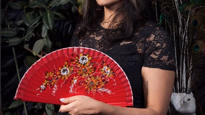 There is a woman sitting in a black dress with lace sleeves. She is holding a red fan with a floral design in front of her chest.