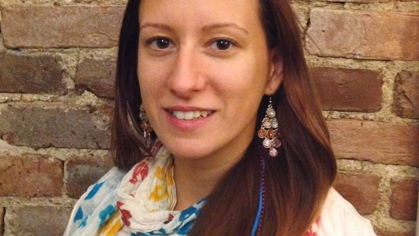 A caucasian brunette female with straight long hair wearing white clothes with colorful accents along with a pair of earrings in front of a brick wall.