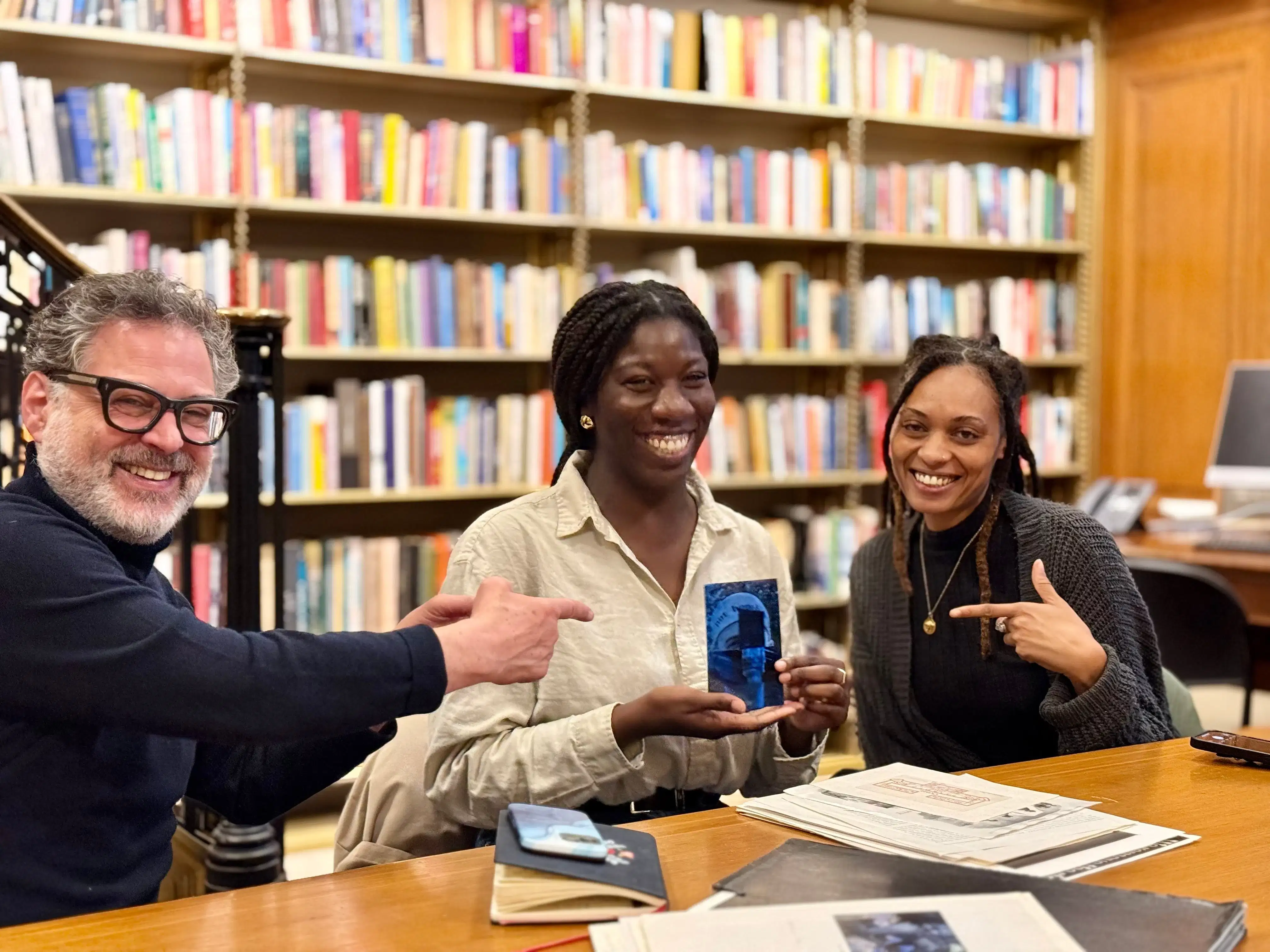 Three people smile at a table in a library reading room, with tall bookshelves behind them; the two on either side point toward the person in the middle, who holds up a small blue booklet.