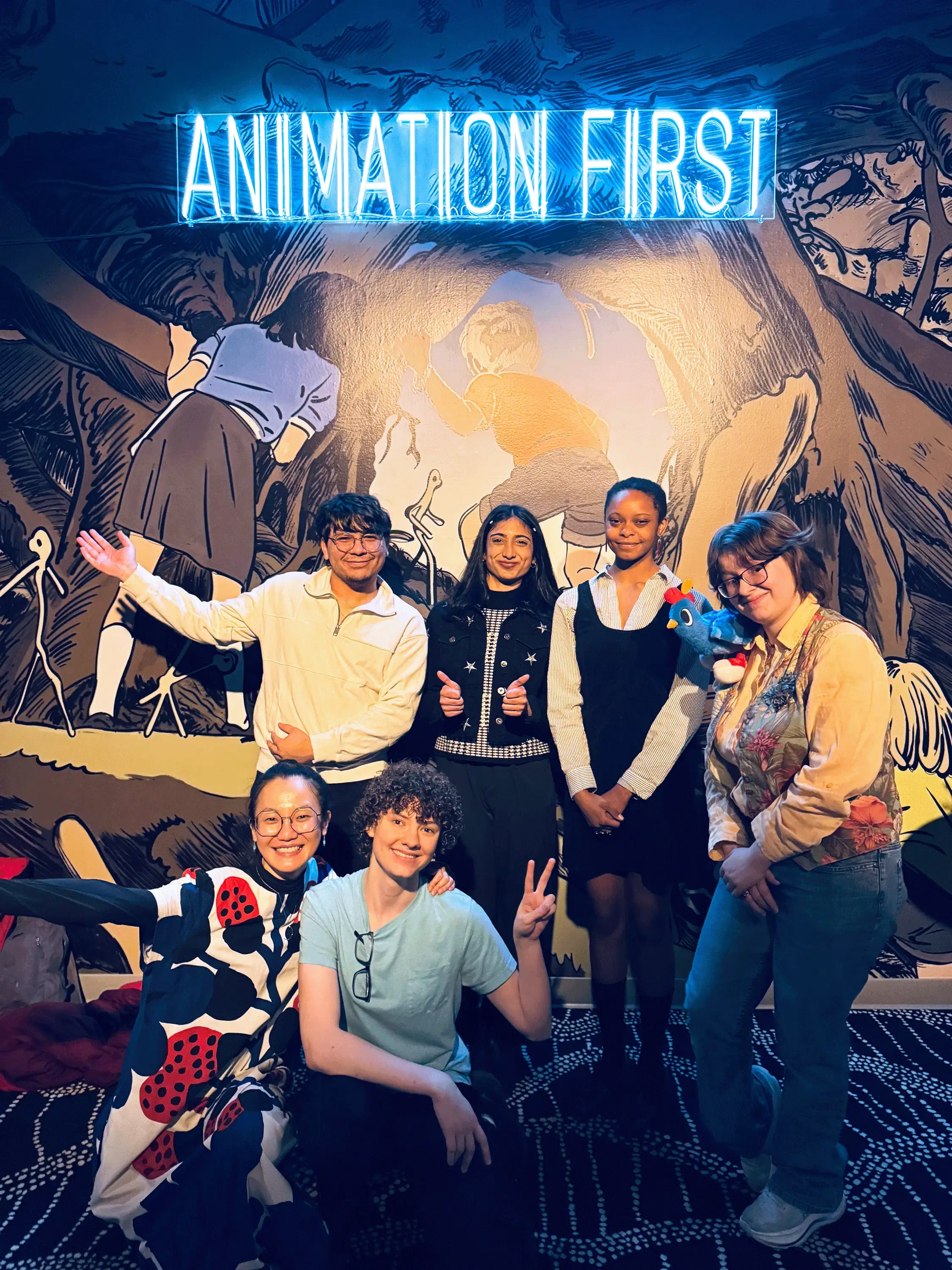 A large group of students and faculty posing for a group photo under a glowing ‘Animation First’ neon sign