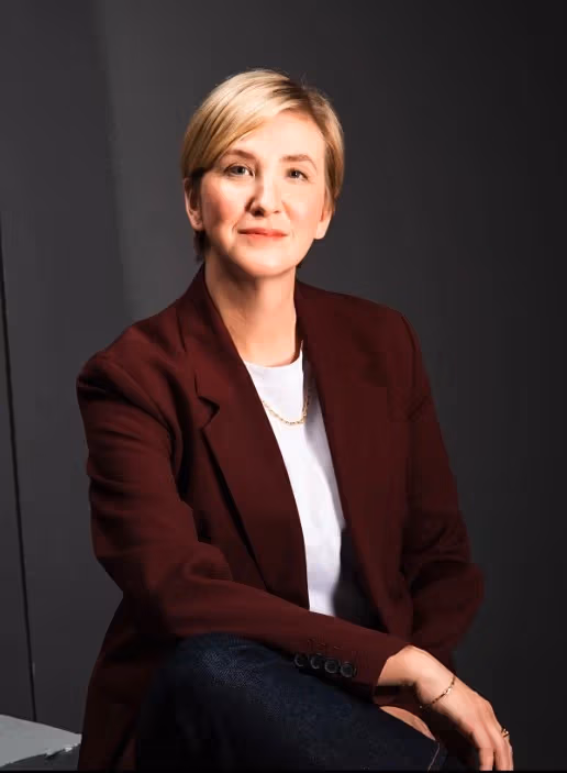 a person with short blonde hair sits in a red blazer