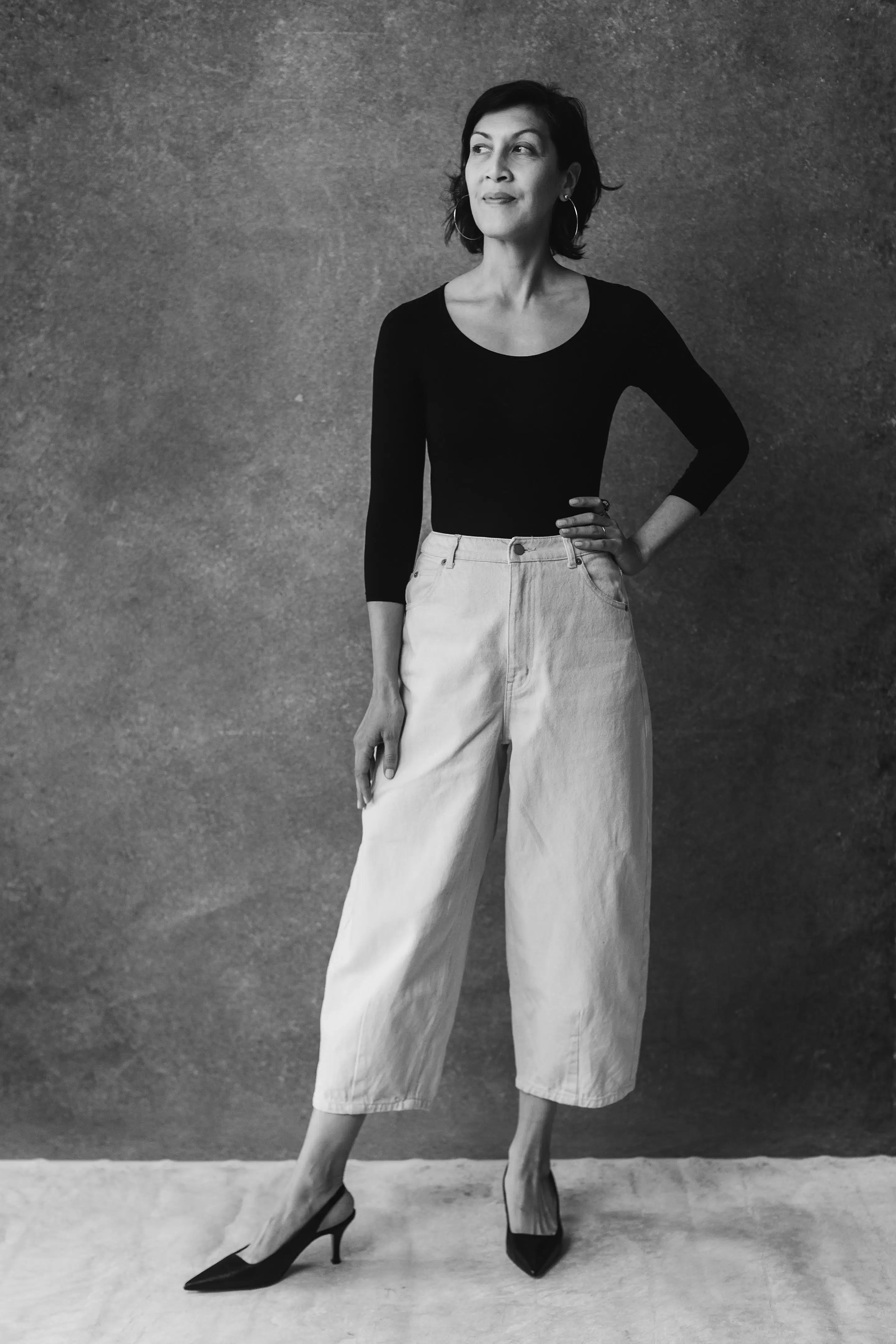 A black and white photo fo a woman standing in front of a gray wall with her hand on her hip and a black scoop neck top on. She is wearing white jeans and black slingback heels.