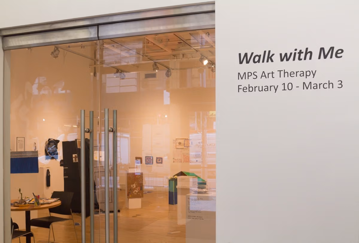 A view from outside of a shop's entrance with transparent glass doors and a write on a wall "Walk with Me-MPS Art therapy February 10-March 3"