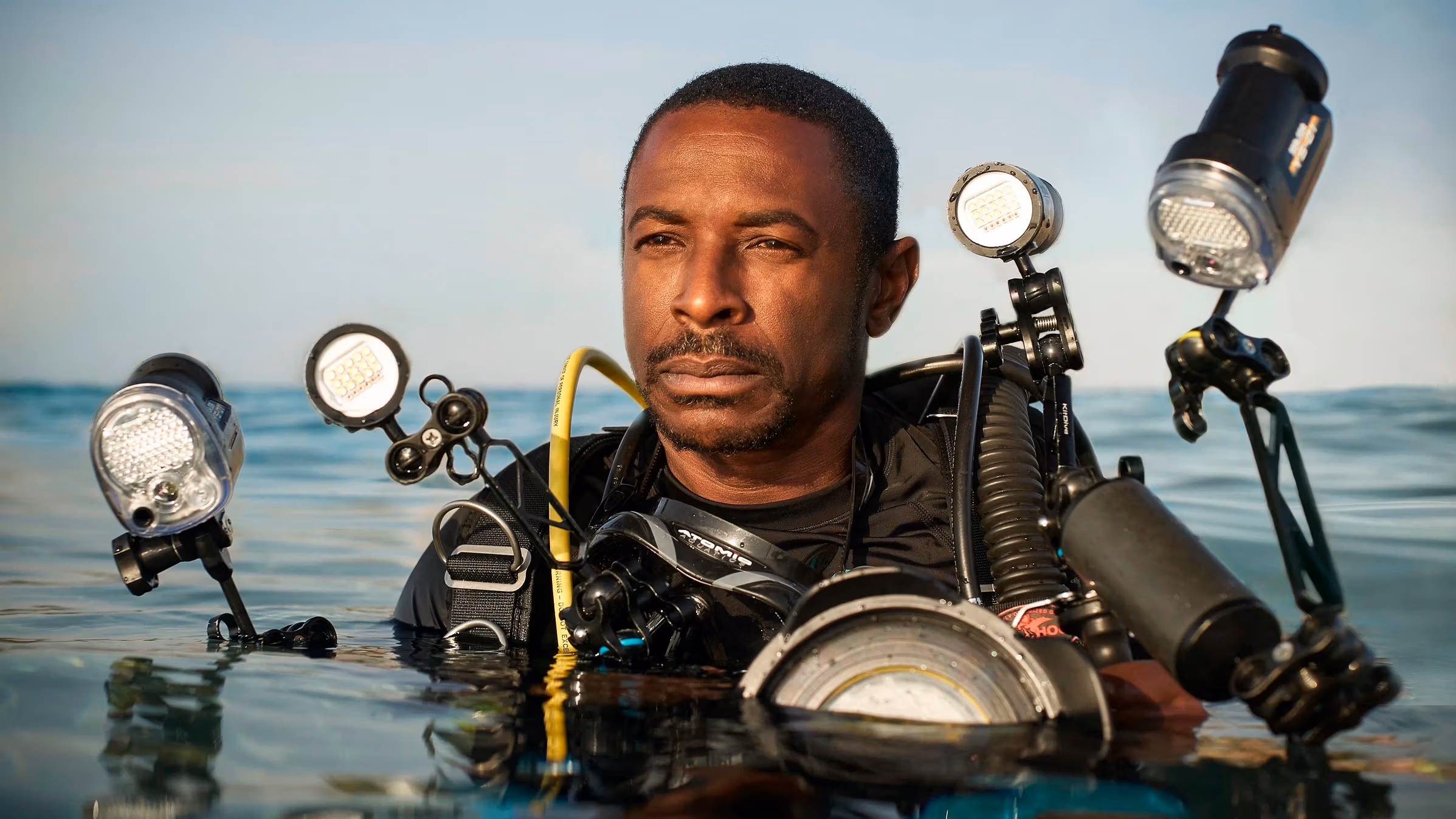 A portrait of a scuba diver in water. He has darker skin and short hair, is wearing a black top, and is equipped with numerous lighting and camera equipments strapped to his body. His body is submerged in water from the chest down.