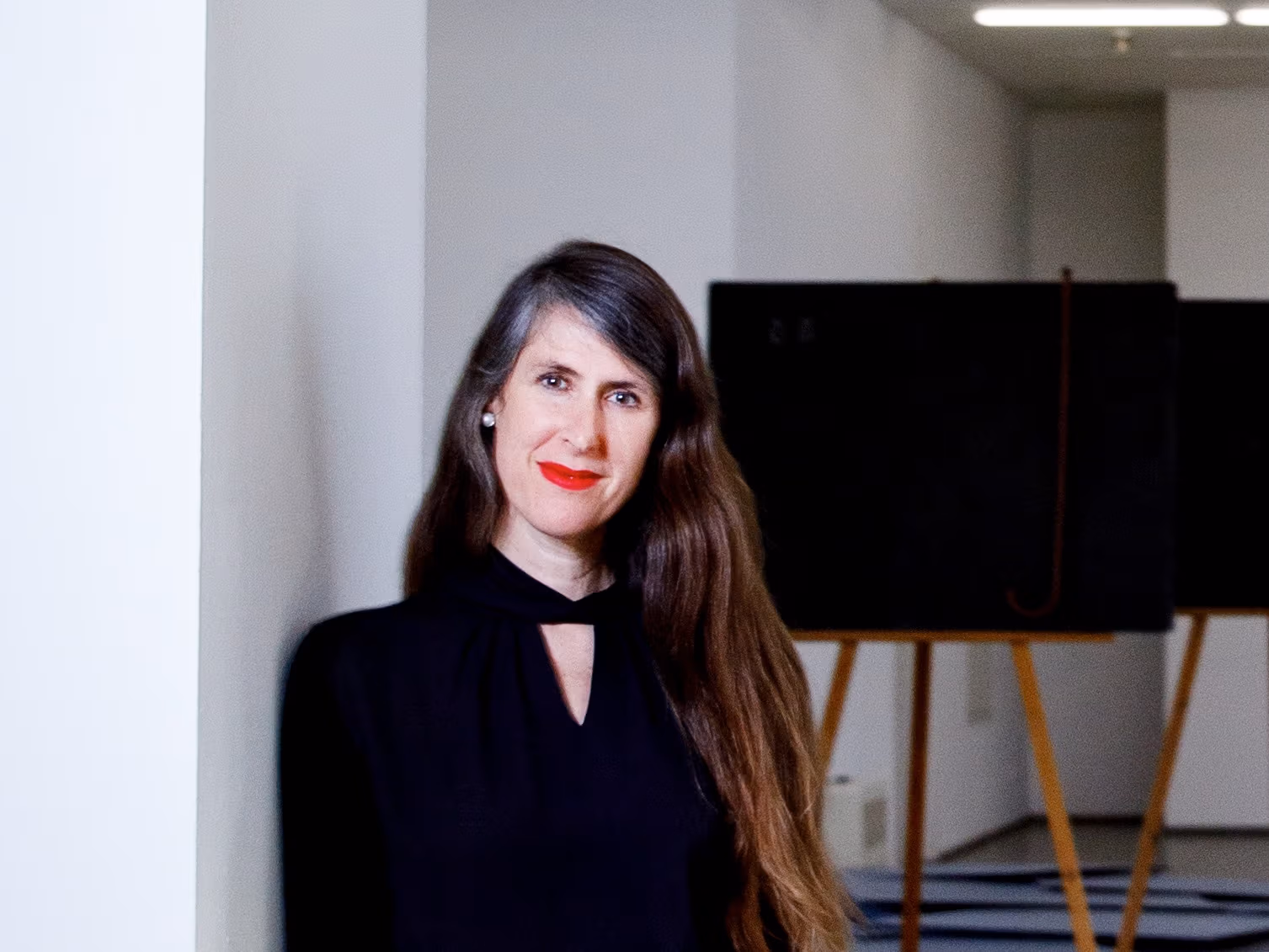 A photograph of a person posed in a sparsely furnished room with bare walls.