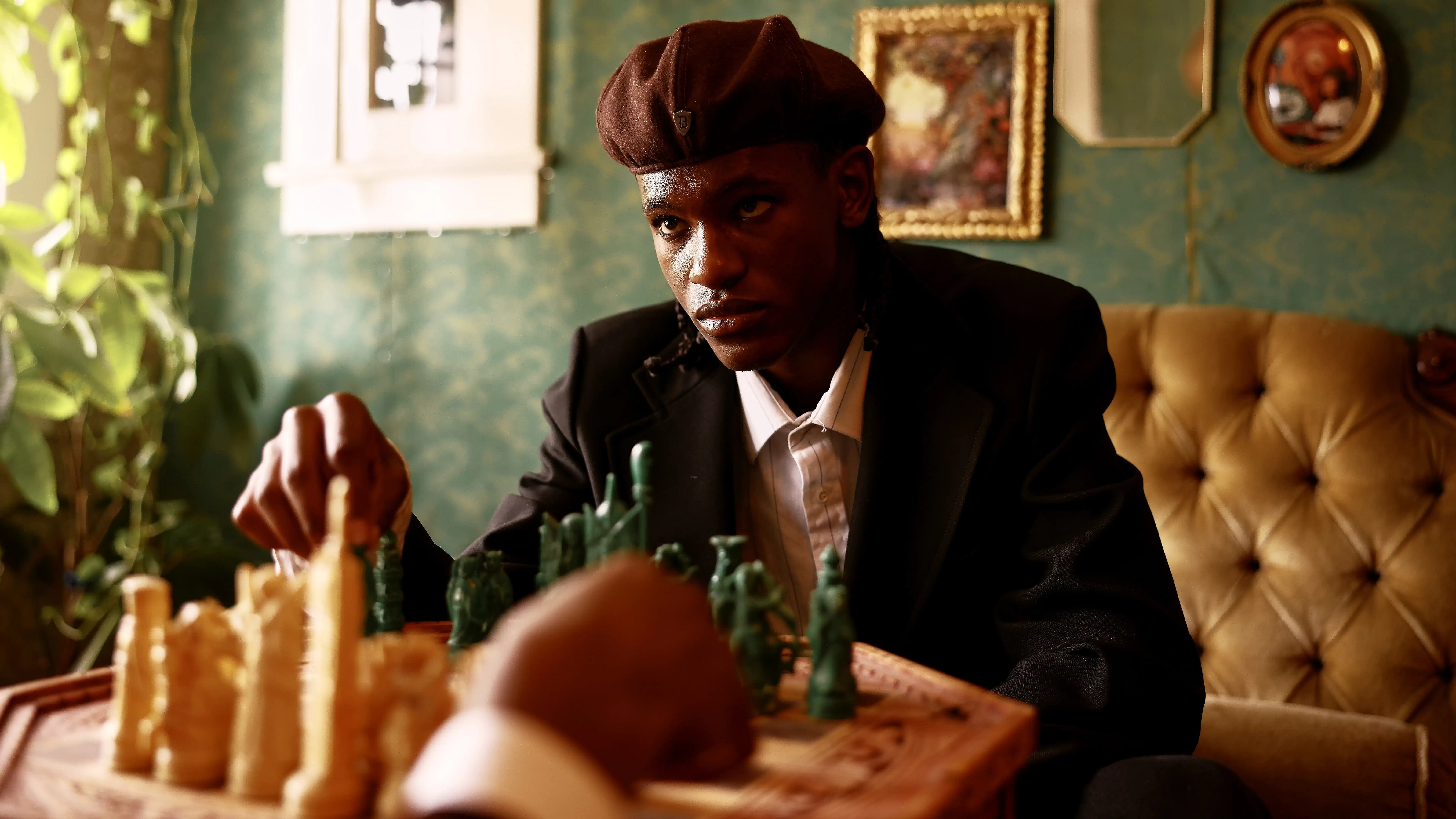 A dark skinned man wearing a beret sits on a velvet couch in a lavishly decorated room, he is bent over a game of chess and he appears to contemplate his next move while making eye contact with an unseen opponent