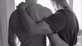 Two men glancing downwards out the window as they hug each other.