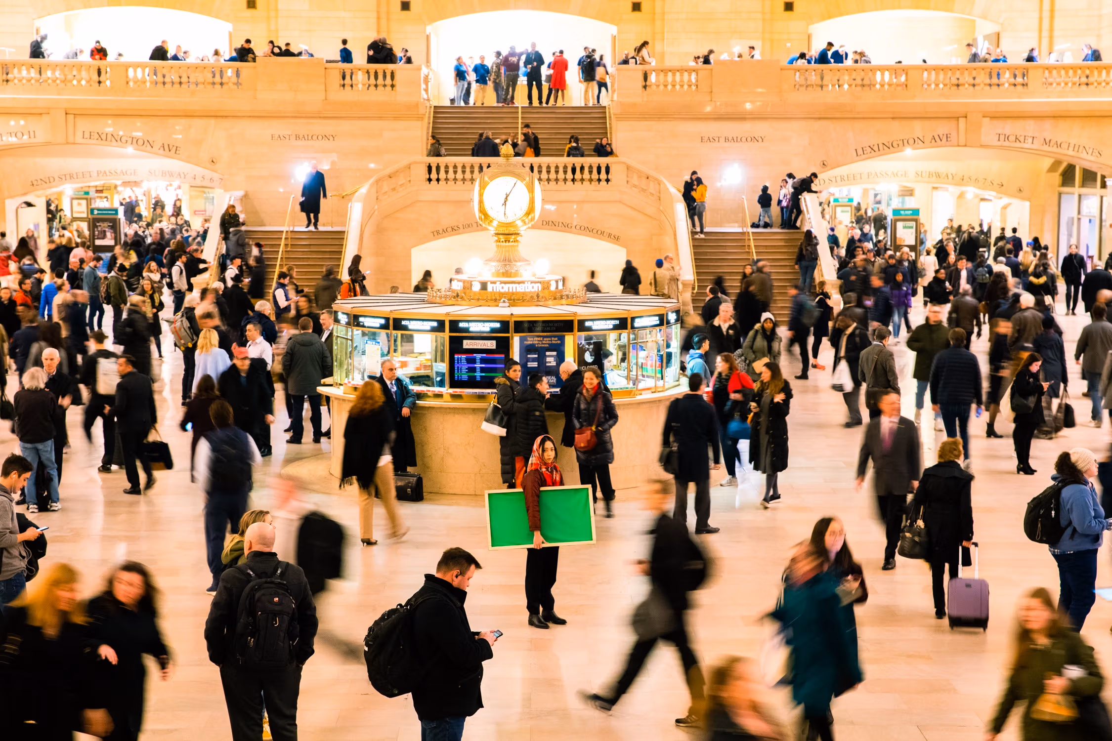 A photo of Grand Central's Main Concourse, with many people bustling around an information booth that's topped with a clock. One person in the middle of the shot is still and in focus, looking at the viewer and holding a bright green canvas under their arm.