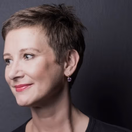 A headshot-style photograph of a person who is posed in front of a blank background, looking off to the left and smiling.