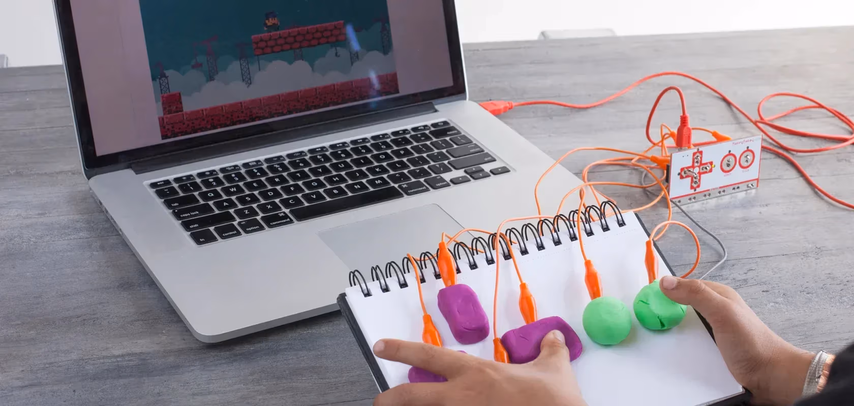 A person holds a notebook with buttons made of colorful molding clay attached by wires to a laptop.