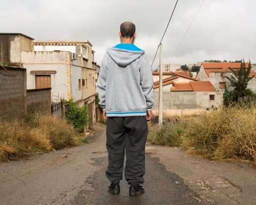 A man with baggy pants and sweatshirt looks down the street in a residential neighborhood of masonry buildings.