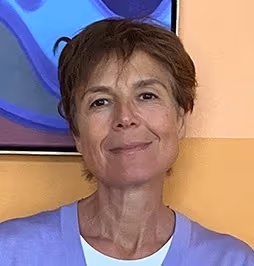 a person with short brown hair and brown eyes smiles in front of a beige wall 