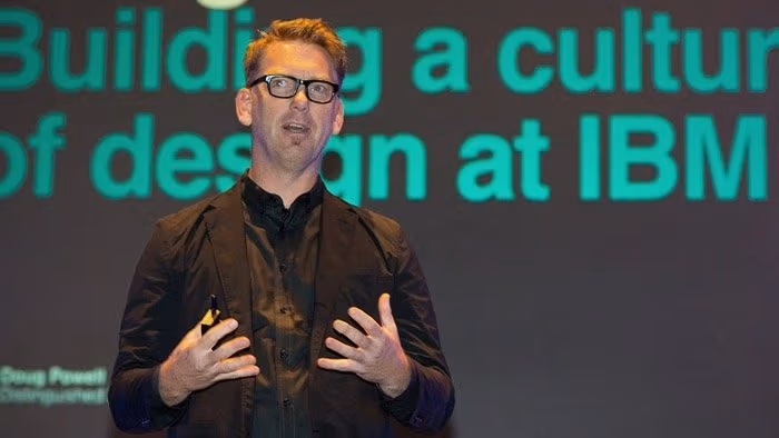 A white man with glasses in a black suit with a black shirt speaking in front of a large screen.