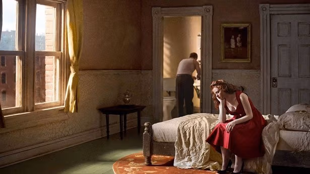 A red haired woman on a bed with a man in brown trousers in the background.