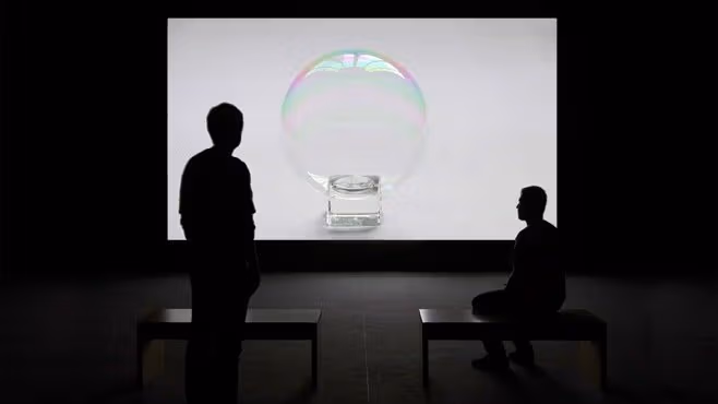 Two people in a room, looking at a screen that has a bubble sitting on a small cube.