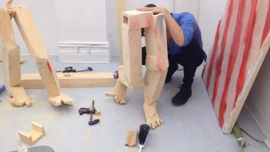 A man working on a wood sculpture of the lower body. Work tools and wood shavings are strewn on the floor.