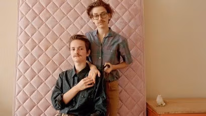 Two brothers both lean up against the mattress in a bedroom.