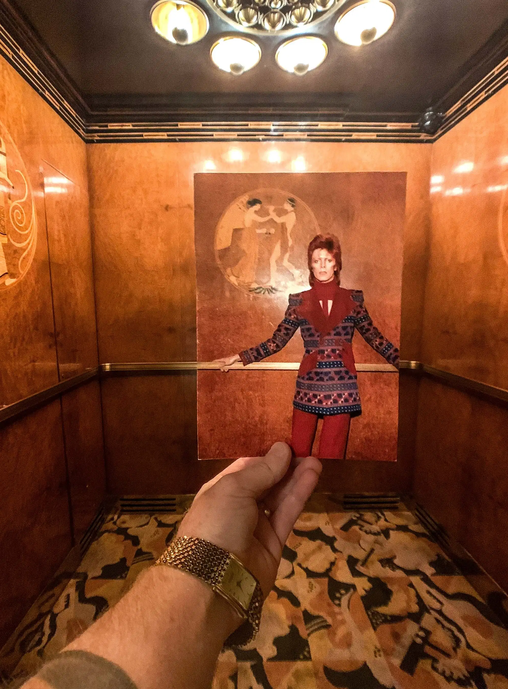 A hand holds a vintage photo in an ornate elevator. The photo features a person in a colorful, patterned outfit matching the elevator's stylish decor.