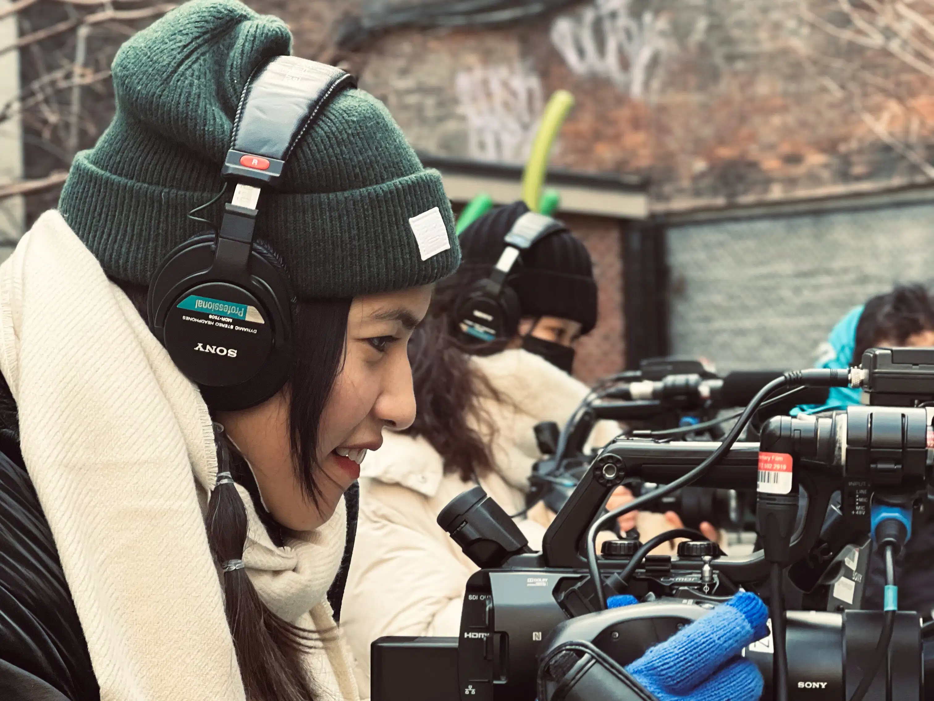 a woman films outside, wearing hat, gloves & headphones and in profile facing right. She looks into her camera. Another woman in a similar position is seen behind her.