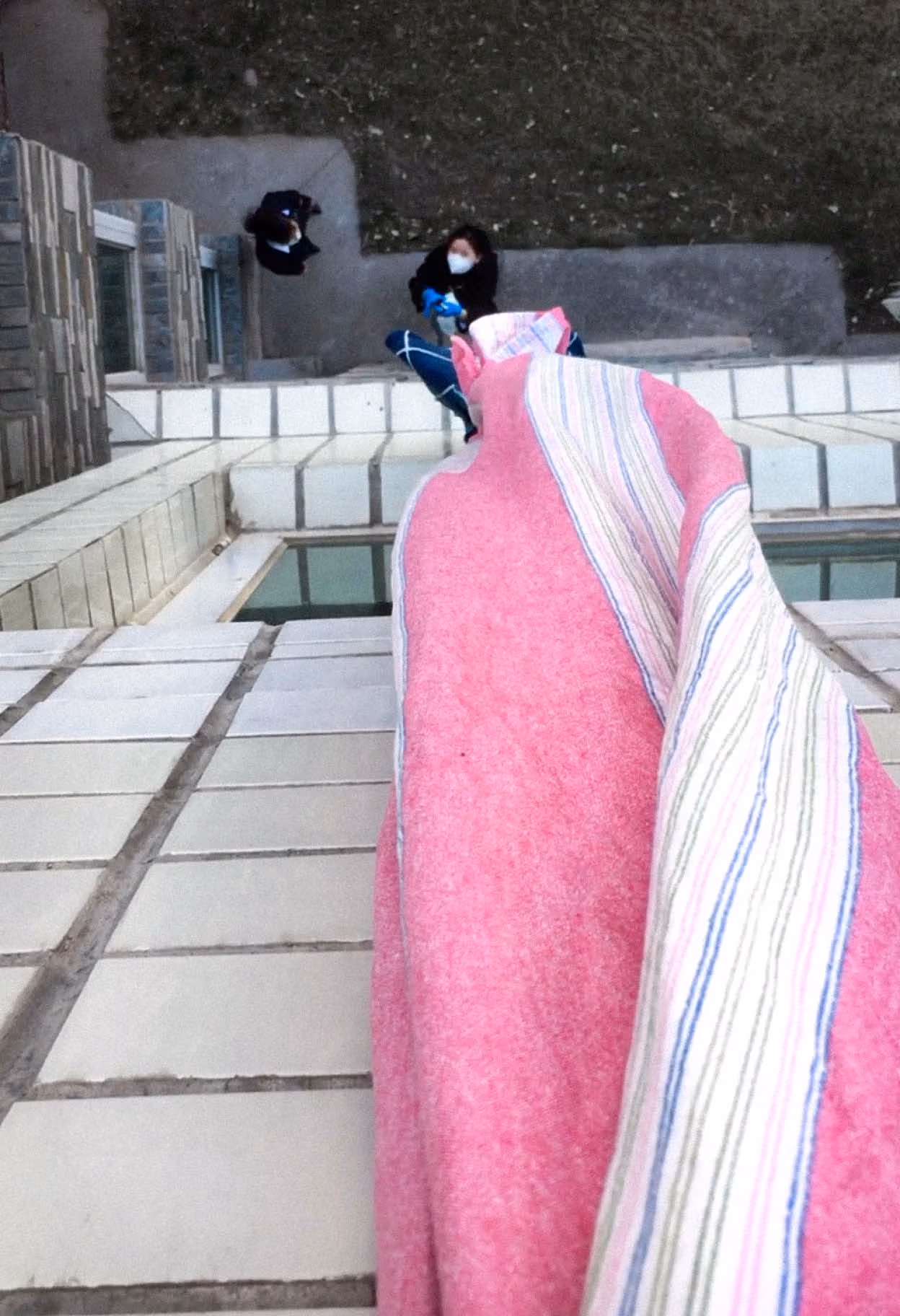 In the photograph, the viewpoint appears to be from above and looks down onto two figures. There is a pink and white stripped fabric that is hanging down the side of the building.