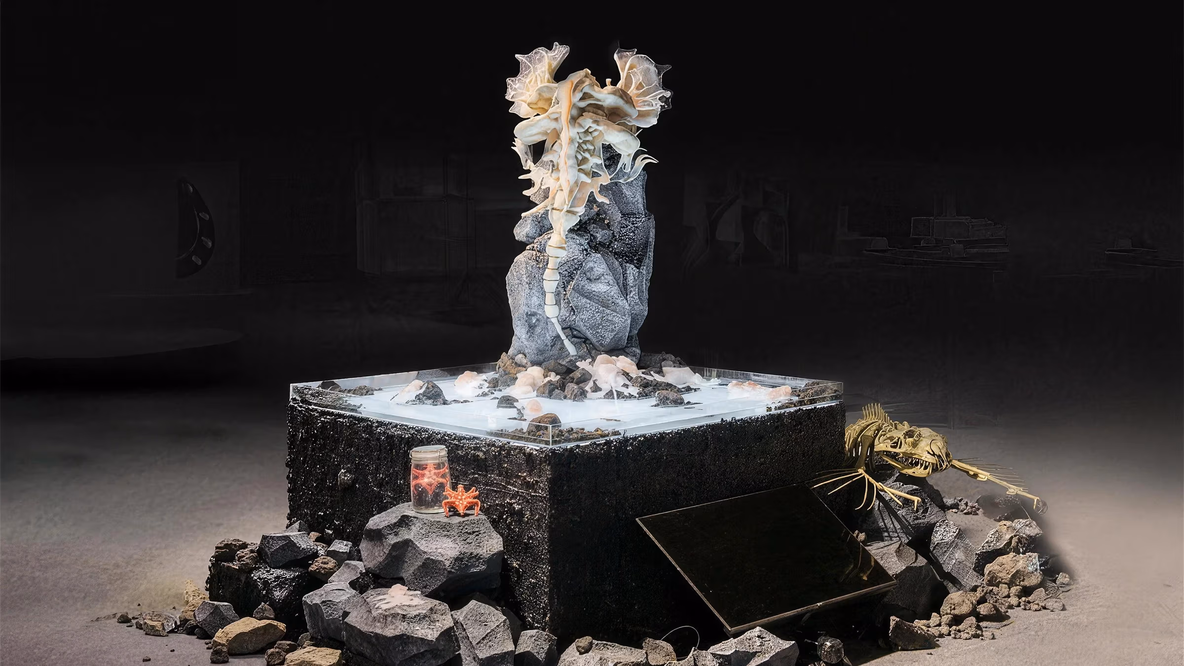 Large sculptural work, showing a silicone sea creature-like form with semi-opaque fins clinging to a large rock-like surface, resting on an illuminated platform surrounded by ocean debris. 