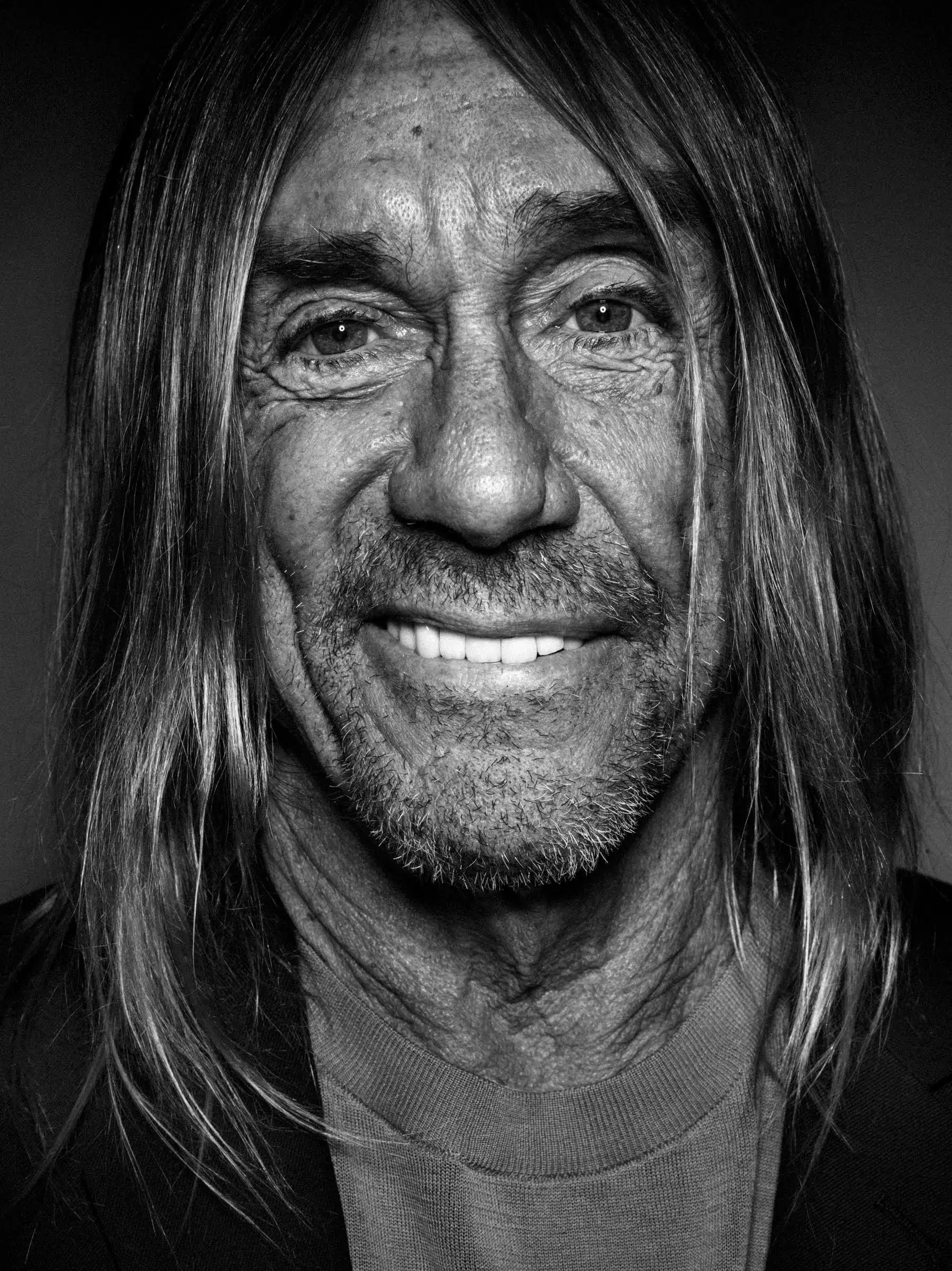 A black and white photo portrait of the singer Iggy Pop, who is looking straight into the camera with a smile. He has long hair and light skin, and is wearing a black cardigan over a gray t-shirt.