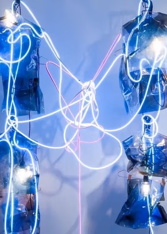 A sculptural installation on a white wall utilizing multiple wires and body-like forms covered in tangled electrical cords.