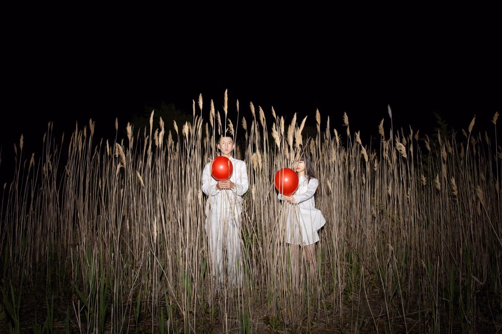 A man and woman wearing all white are each holding a red balloon, standing in tall reeds at night.