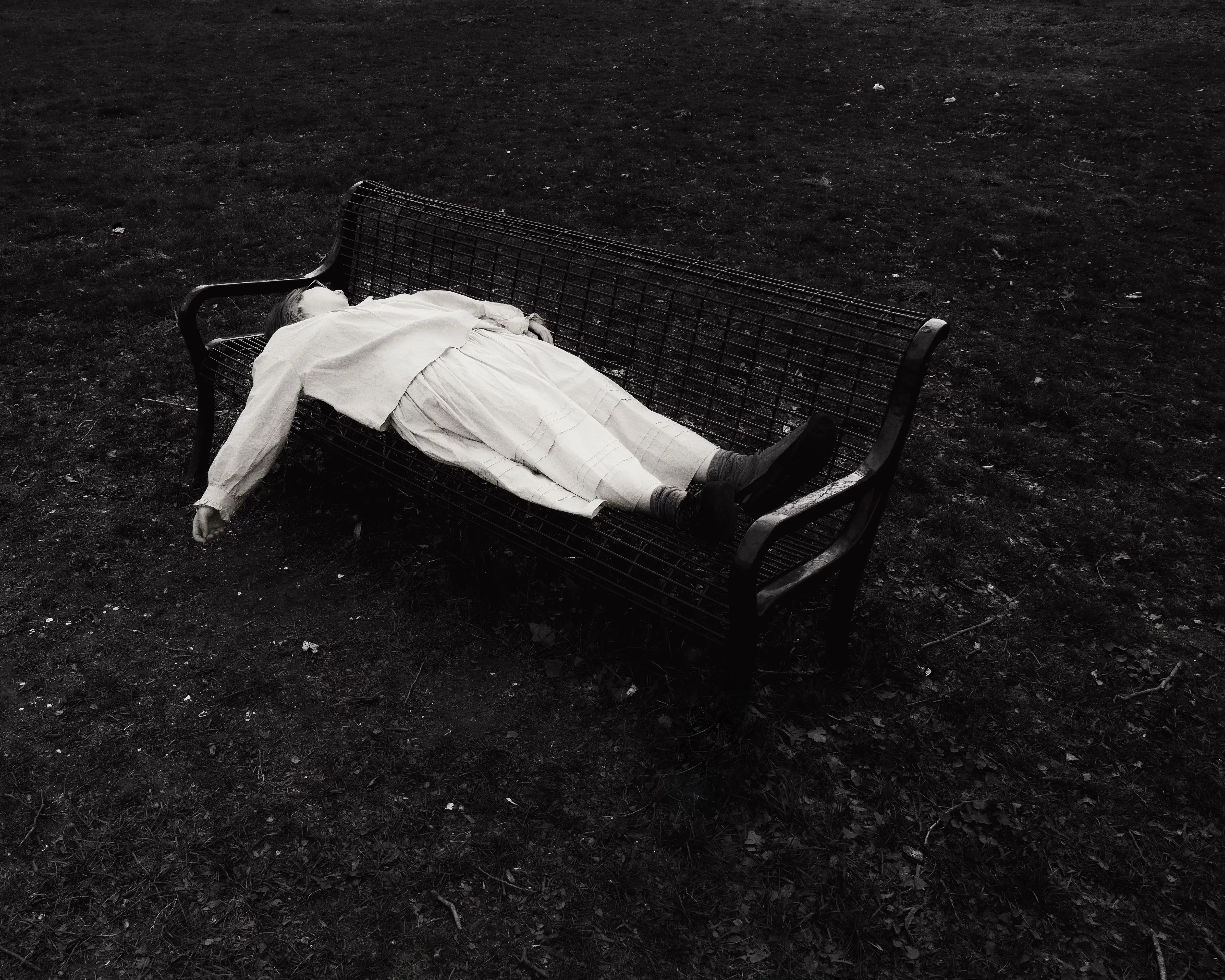 A black and white photograph of a girl lying on a bench, her hand hanging loosely, face obscured. The bench and her body are surrounded by chaotic bundles of grass.