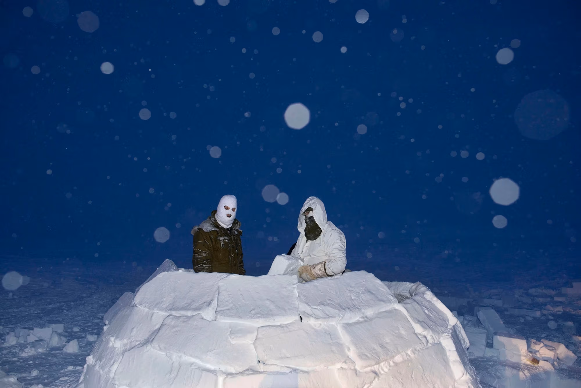A color photograph of Two soldiers, wearing masks in an igloo made of snow blocks, at dusk while it snows, lit with flash.