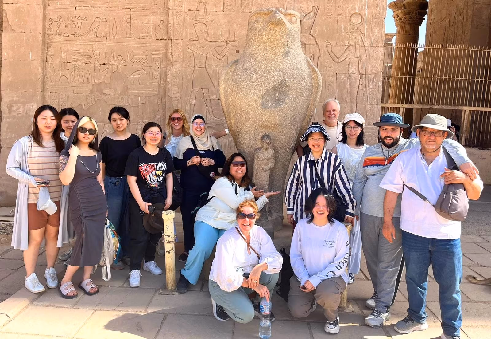 A group of students and faculty members, some wearing sunglasses and hats stand and pose in front of a ancient building in Egypt. 