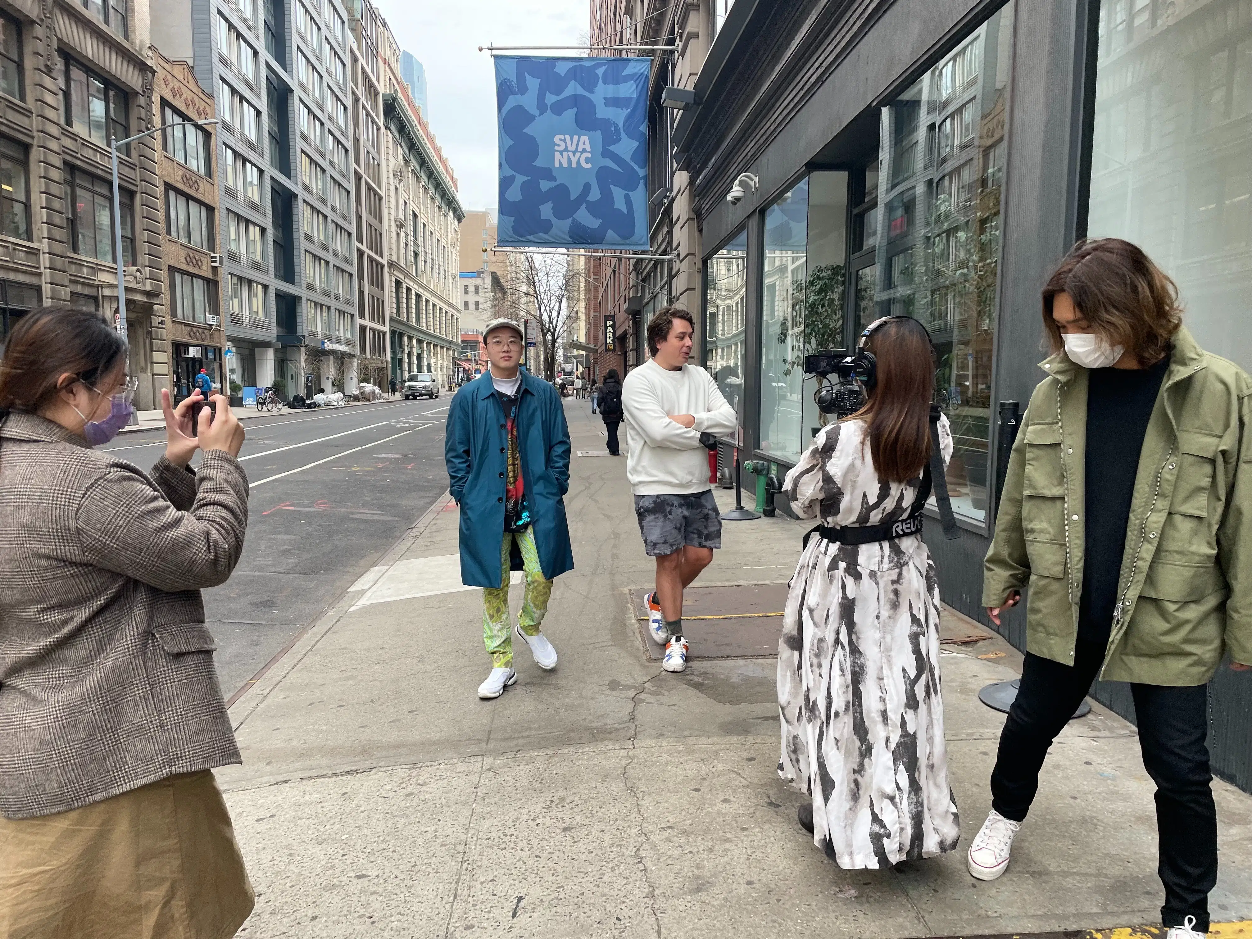 5 students walk towards the camera, 2 walk backwards holding cameras, filming another two. A blue SVA NYC banner flies in the background
