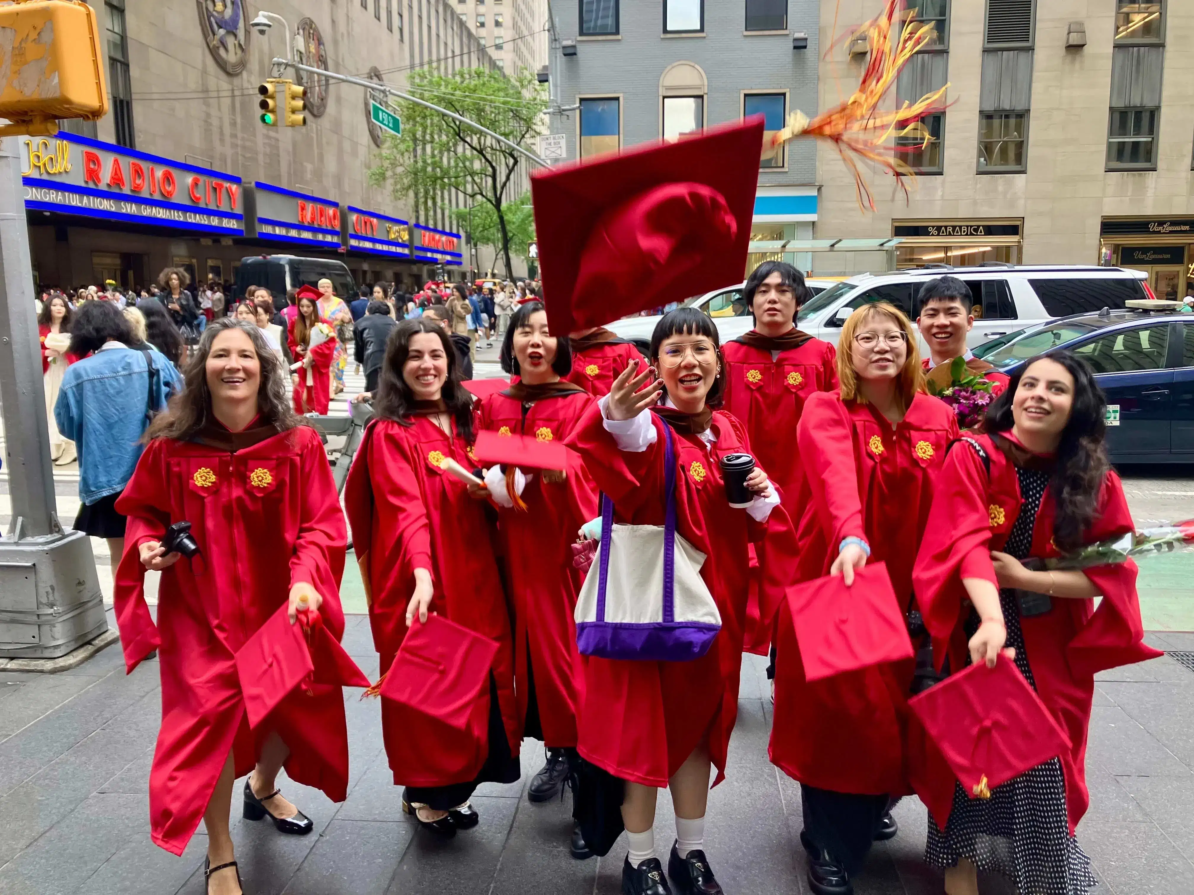The Masters in Digital Photography graduating class of 2025, in the iconic SVA red graduation gowns, in front of the famed Radio City Music Hall where the commencement ceremony just took place. The students are throwing their caps in excitement. 