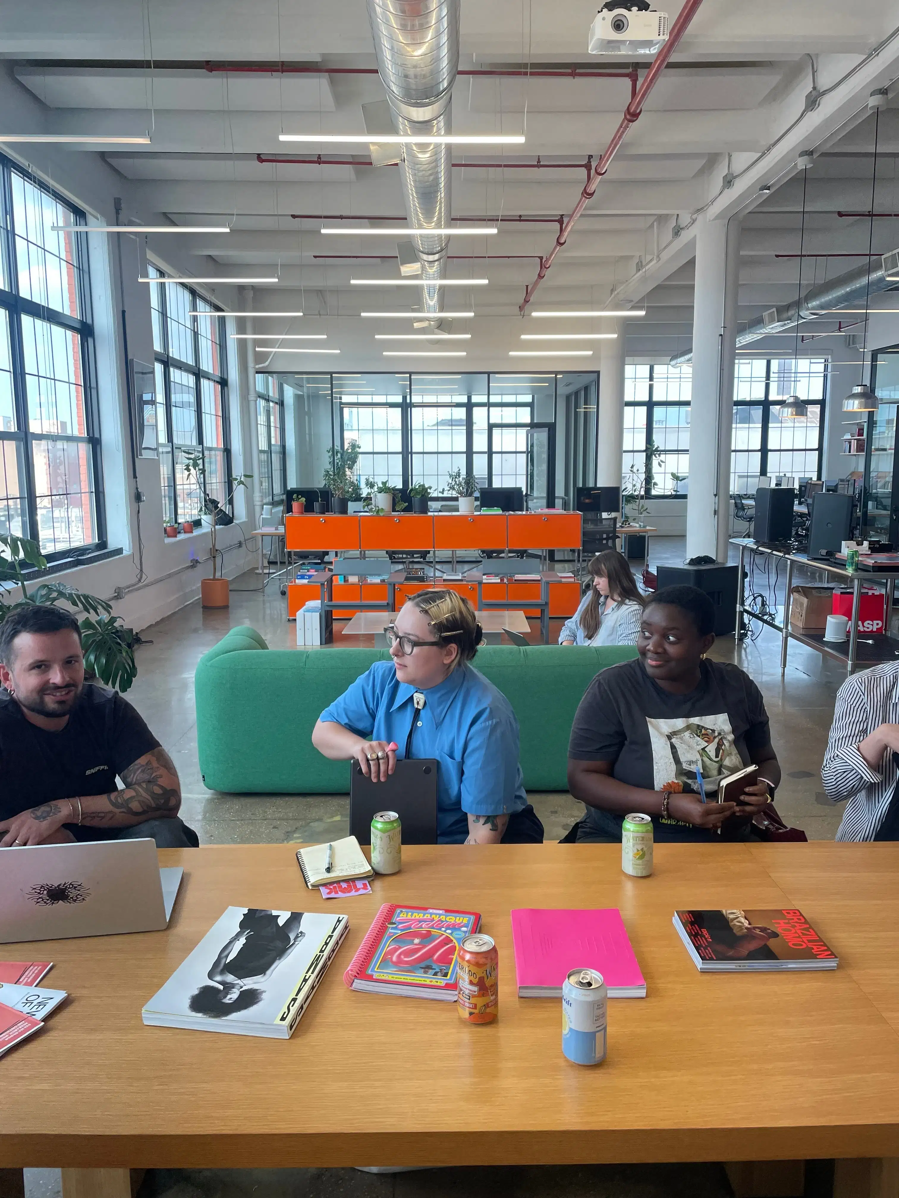 Alt text: Wide photo of a bright, industrial loft workspace with tall grid windows and exposed ducts. In the foreground, three people sit at a long wooden table—one with a laptop, others with notebooks and books—while soda cans and colorful magazines are spread across the tabletop. A green sofa and rows of desks and computers fill the background.