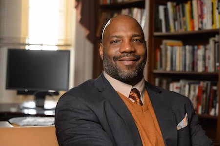 A portrait photograph of writer and academic Jelani Cobb sitting in an office with a desk and bookcase behind him.