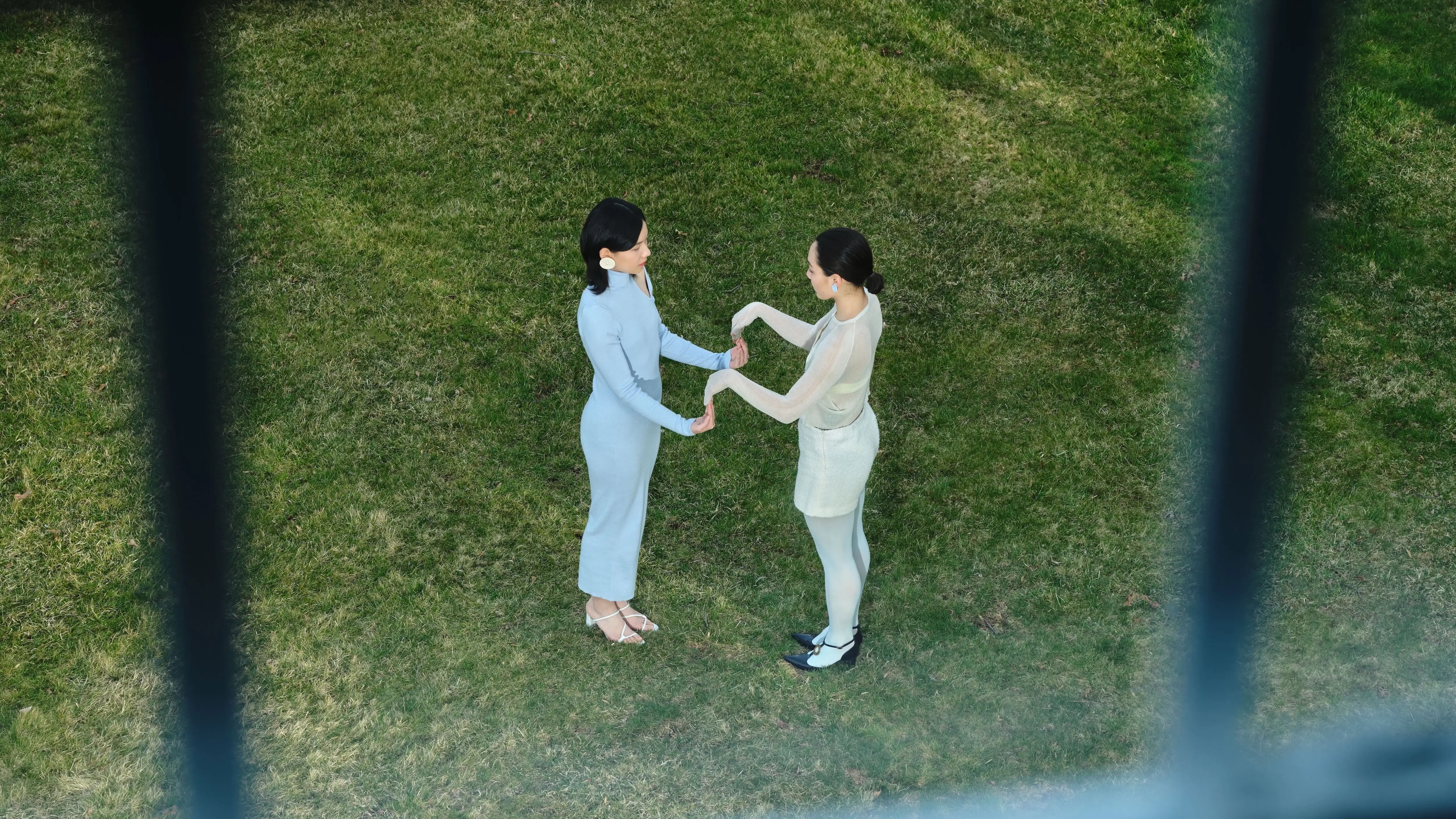  Two women stand face-to-face on a green lawn, gently holding each other’s hands, seen through a window grid from above.