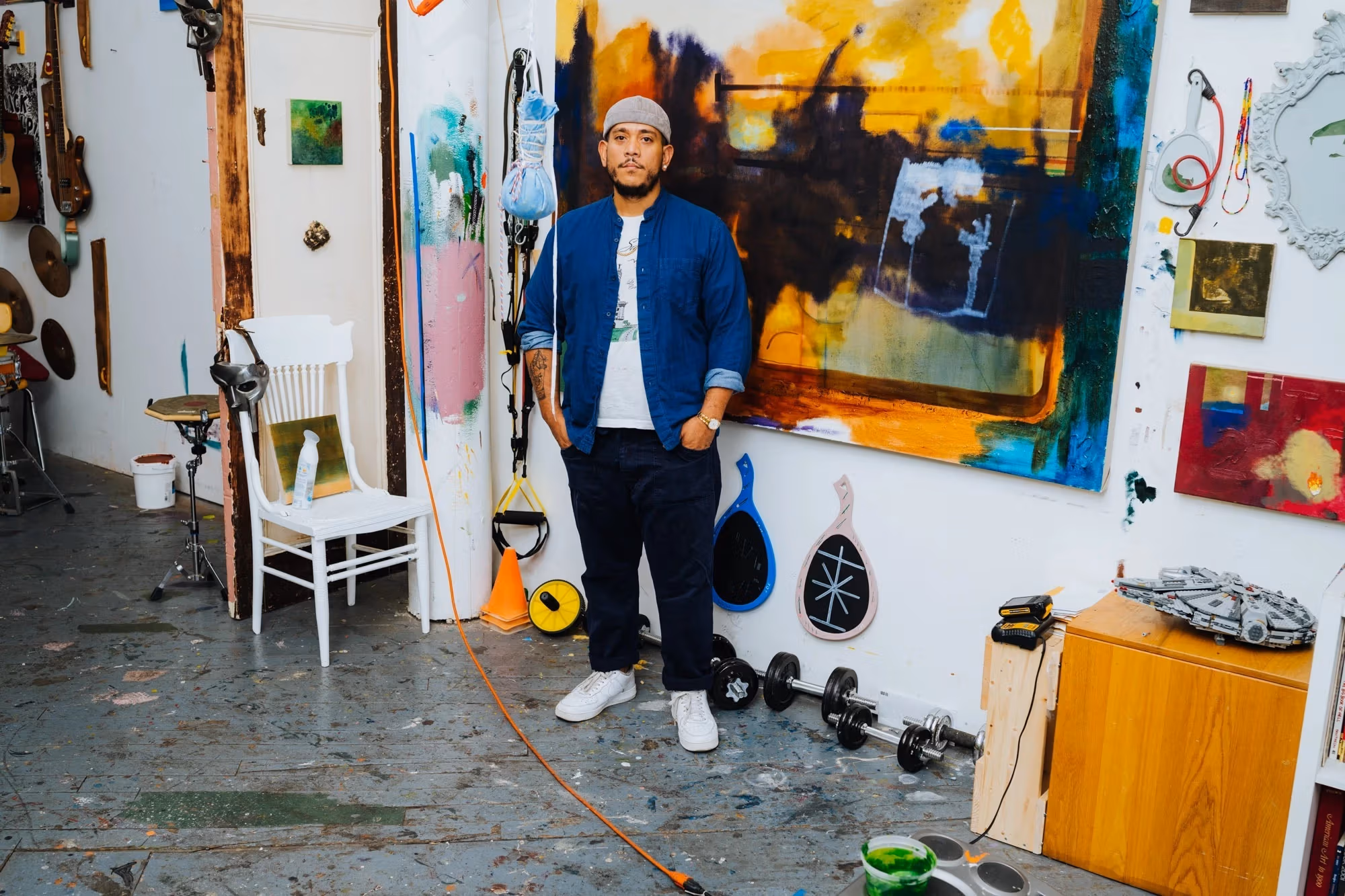A photograph of Kenny Rivero in his artist studio. Kenny is standing in front of a wall with a paintings and other objects hanging on the wall. Kenny wears a white shirt with a blue button-down over it and black pants. His hands are in his pocket and he is looking at the camera.