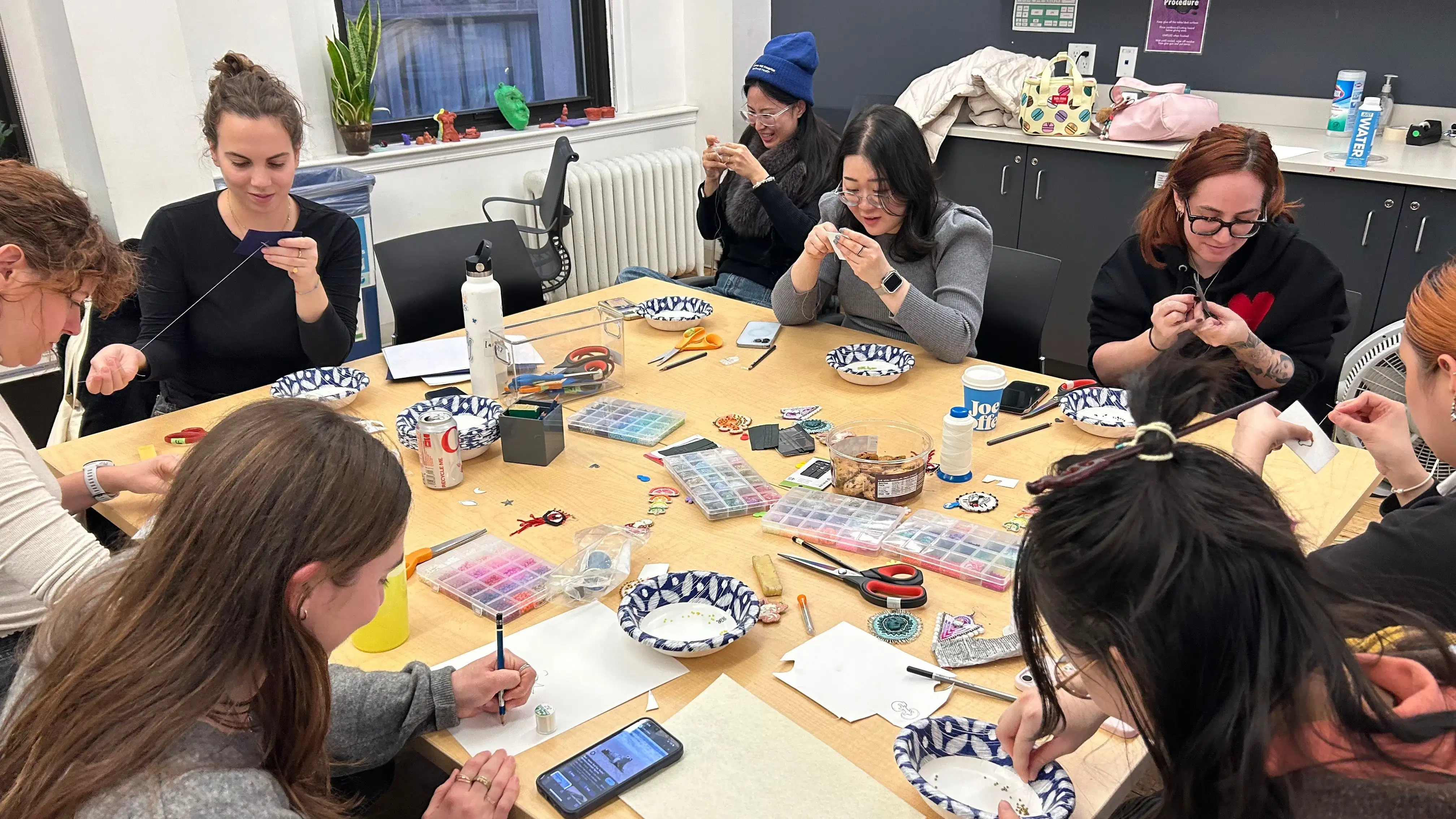 students gathered around a square wood table covered in art supplies working on a beading project 