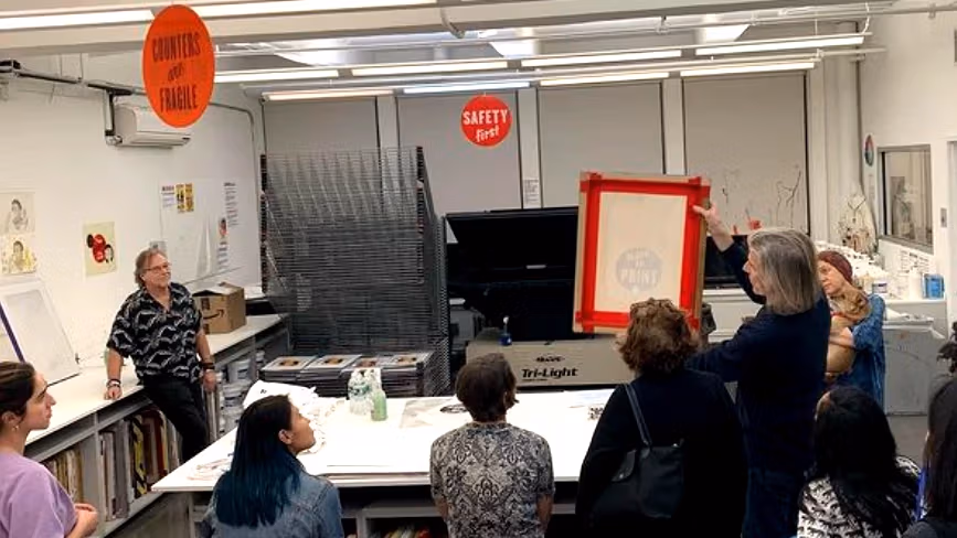 A group of people in the SVA printshop talking in a circle. Someone is showing the group a prepared screen printing screen. 
