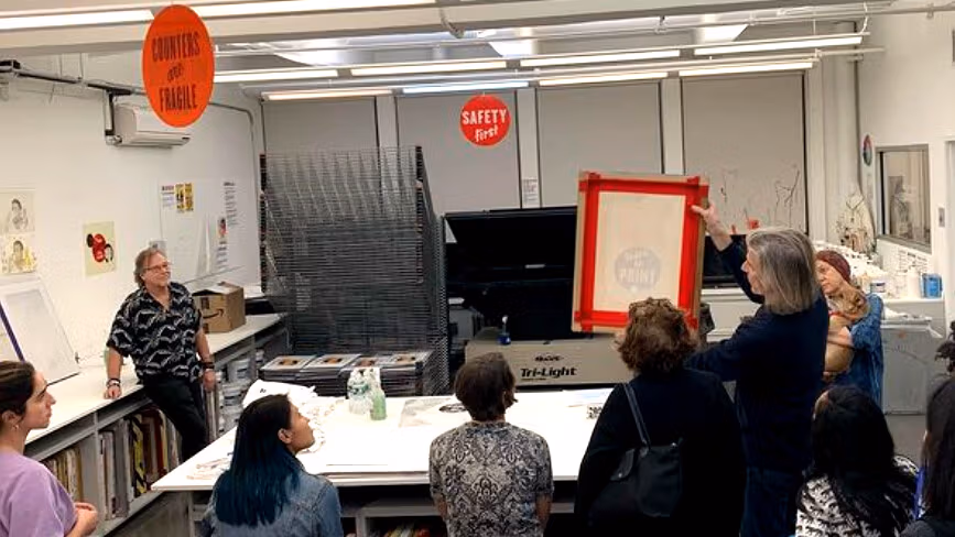 A group of people in the SVA printshop talking in a circle. Someone is showing the group a prepared screen printing screen. 