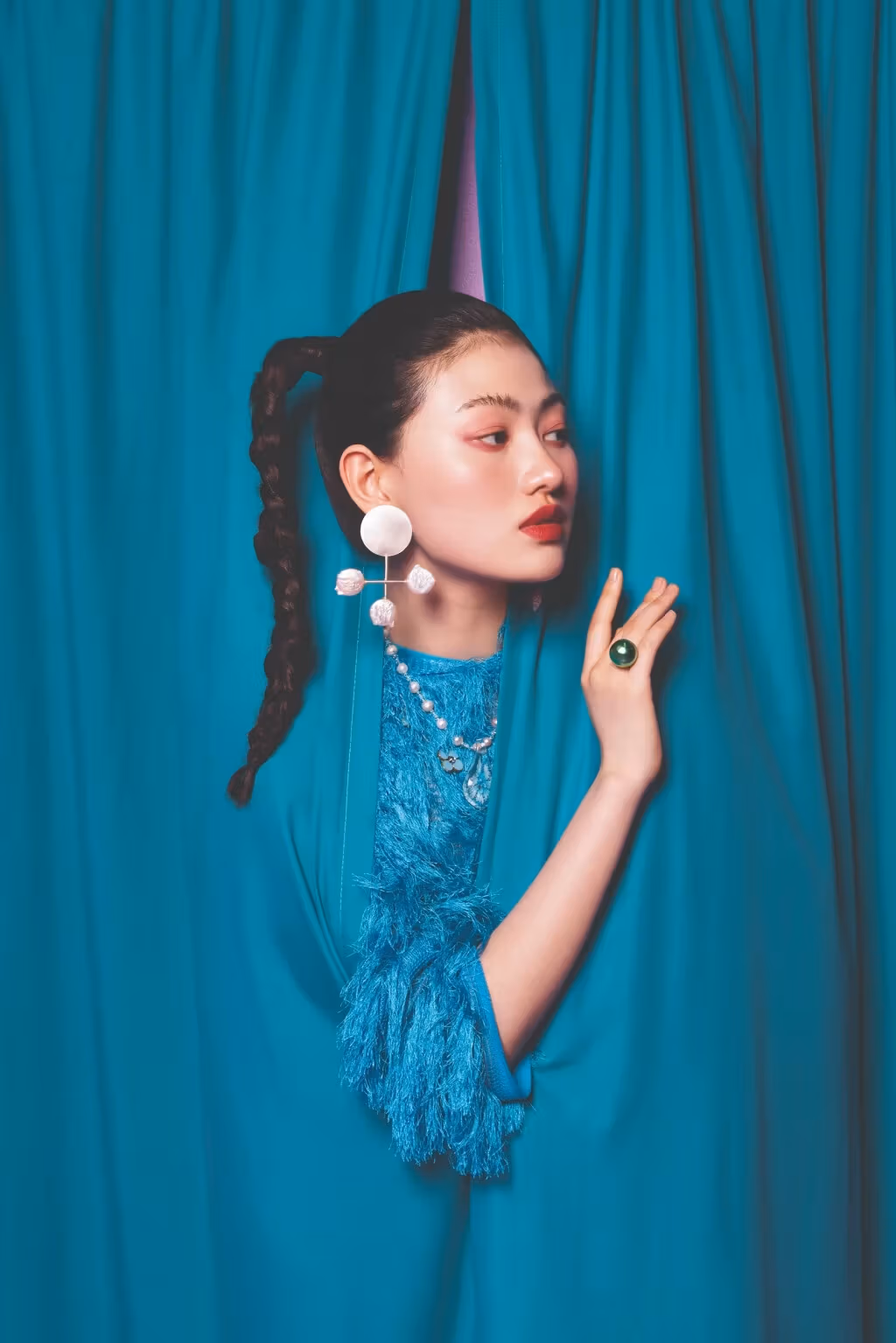 A woman peers out from behind a set of blue curtains. She is wearing a large white earring and a round green ring