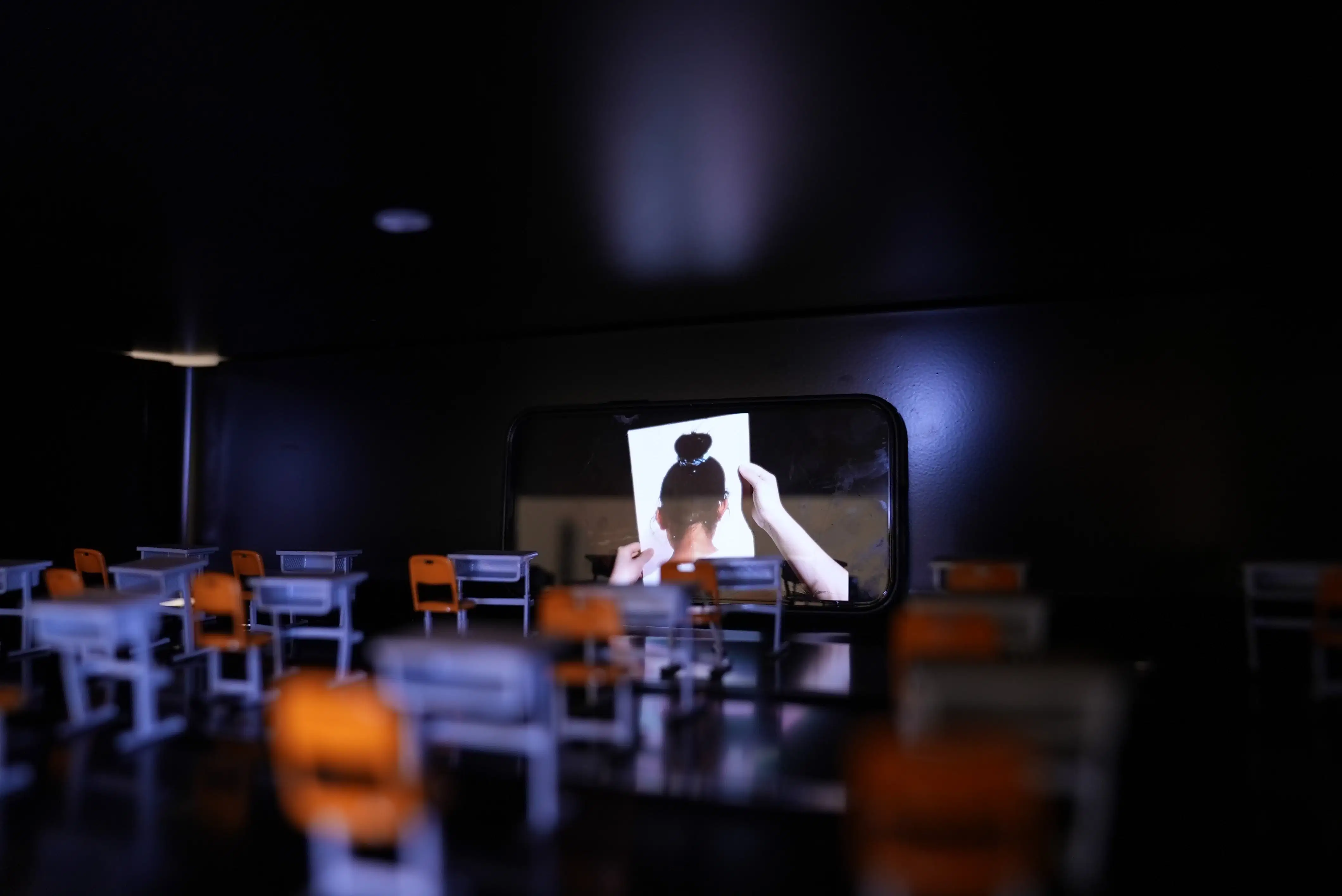 A video of a little girl reading an apology letter is inside the school’s desk. Darkly lit image and still from a video installation.