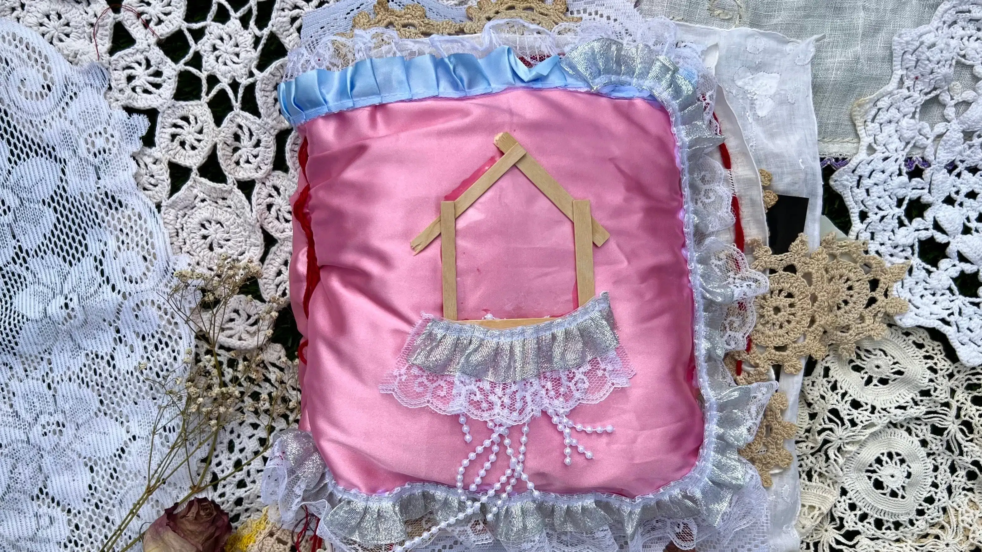  soft pink satin square pillow book cover with image of house made of light wooden sticks with thick strip of white lace and thin strips of threaded beads hanging down. Blue lace is sewn into the top of the cover. A film strip is seen at the bottom of the pillow. This pillow book is placed on pieces of white and off-white antique like lace.