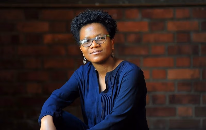 a person in a blue sweater sits in front of a brick wall and smiles at the camera 