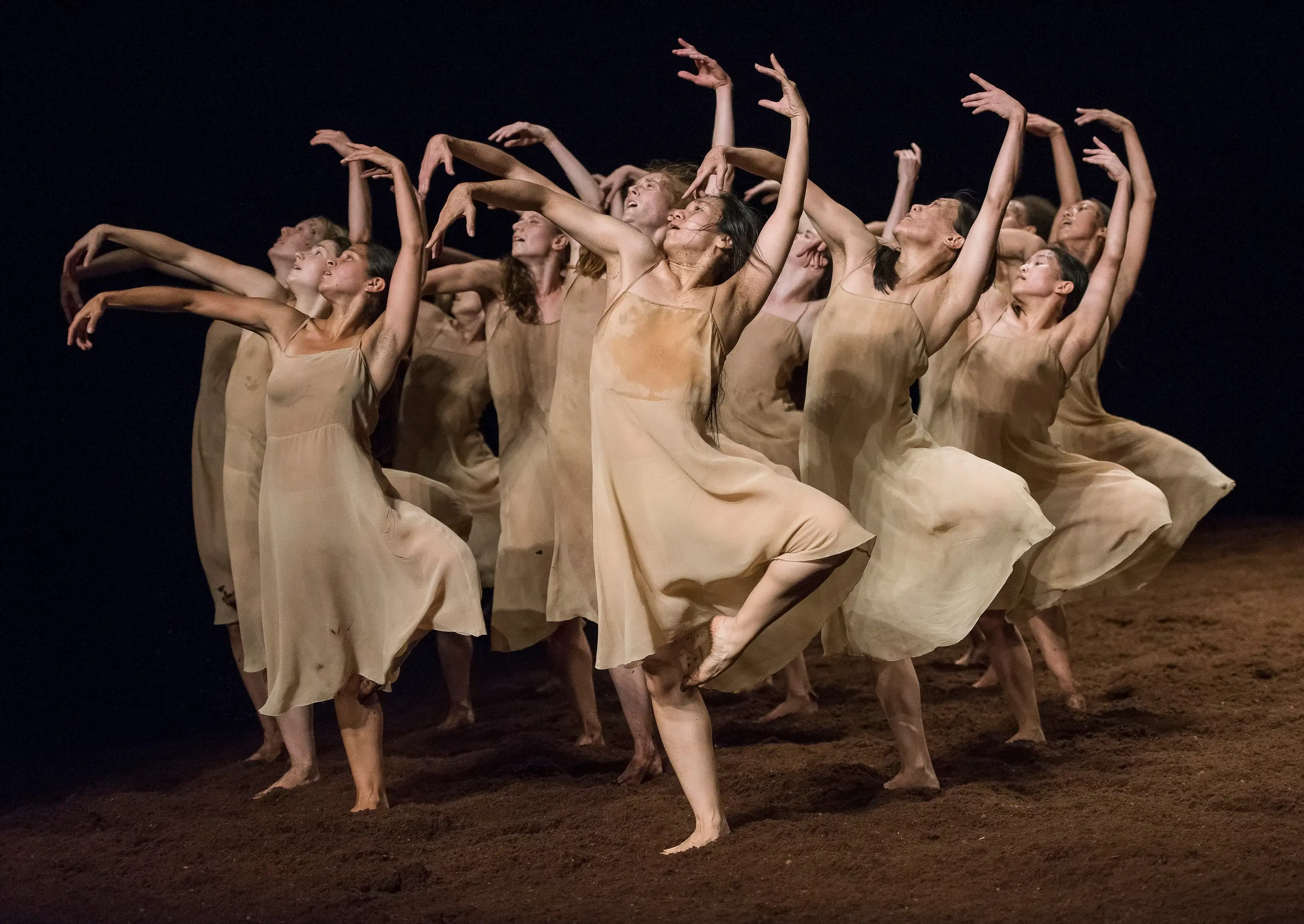A group of dancers in beige dresses on a dirt floor standing on one leg with the other leg bent, holding their hand above their heads while looking up and to the left.